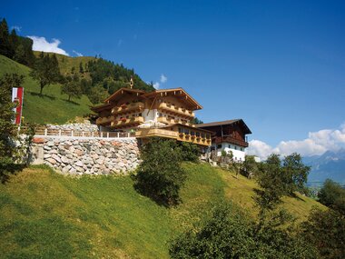 A traditional mountain chalet on a green sunny hillside with blue sky in the background. | © Pfefferbauer Zell am See
