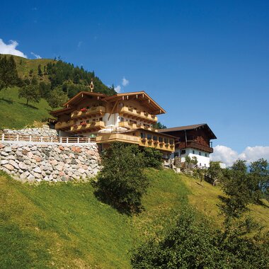 A traditional mountain chalet on a green sunny hillside with blue sky in the background. | © Pfefferbauer Zell am See