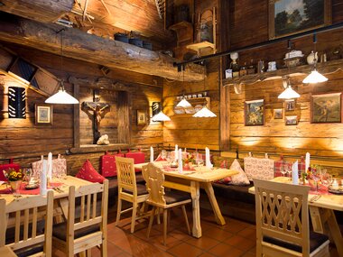 Cozy interior of a hut with timber walls, tables with candles, colorful cushions, and decorative pictures. | © Dorfstadl Kaprun