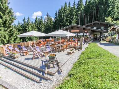 Outdoor of a mountain hut with sun loungers, tables, and umbrellas amidst a dense forest. | © Enzianhütte Zell am See