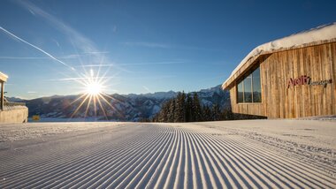 Snow-covered slope with a modern cabin in a ski area, sunrise over the mountains. | © Schmittenhöhe