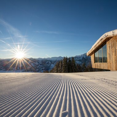 Snow-covered slope with a modern cabin in a ski area, sunrise over the mountains. | © Schmittenhöhe