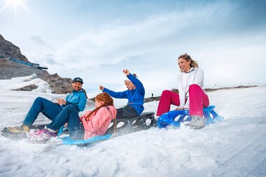 Group of people sitting and laughing on a sled in the snow on a mountain sunny weather. | © Kitzsteinhorn