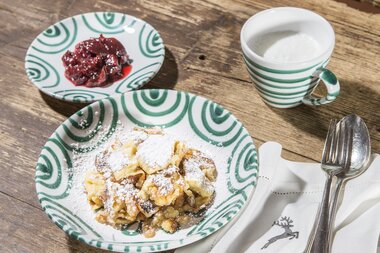 Kaiserschmarrn dusted with powdered sugar and coconut flakes, served with lingonberry jam and a milk drink on a wooden tray. | © Nikolaus Faistauer Photography
