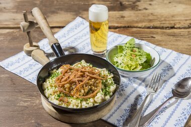 Cheese dumplings with fried onions and roasted meat, served with salad and a beer on a wooden table. | © Nikolaus Faistauer Photography