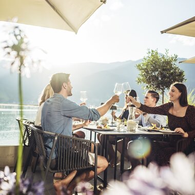 People enjoy dinner with a view of Lake Zell on a sunny day, under umbrellas, while drinking wine. | © Zell am See-Kaprun Tourismus