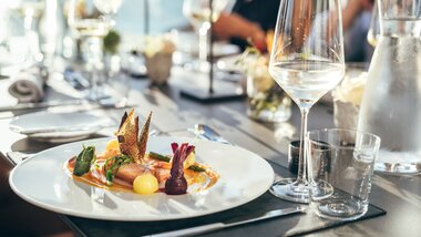 Elegant restaurant table setting with a gourmet dish on a white plate, surrounded by wine glasses and water carafes. | © Zell am See-Kaprun Tourismus
