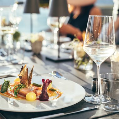 Elegant restaurant table setting with a gourmet dish on a white plate, surrounded by wine glasses and water carafes. | © Zell am See-Kaprun Tourismus