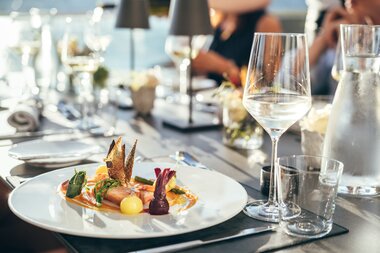 Elegant restaurant table setting with a gourmet dish on a white plate, surrounded by wine glasses and water carafes. | © Zell am See-Kaprun Tourismus