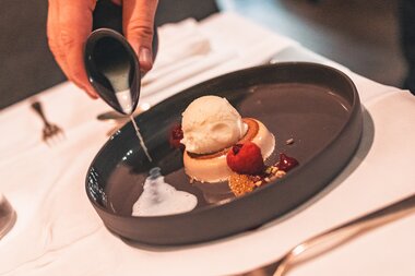 A dessert with a scoop of ice cream, fresh berries, and a subtle sauce on a black plate, served on a white tablecloth. | © Johannes Radlwimmer
