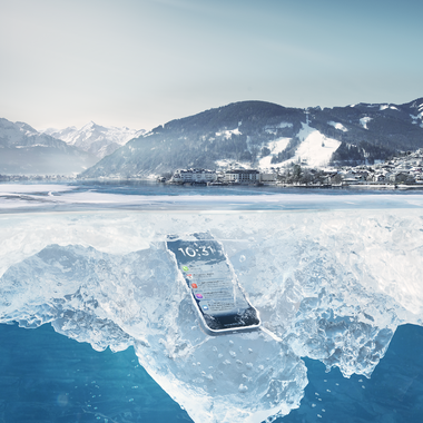 A smartphone is embedded in an ice layer on a frozen lake, surrounded by snow-covered mountains and a winter landscape background. | © Zell am See-Kaprun