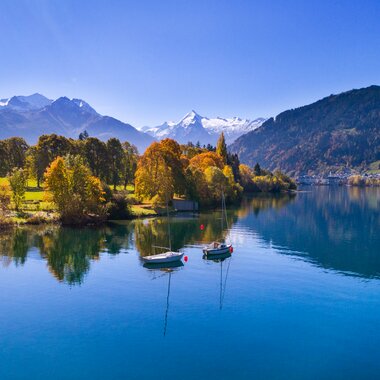 View of a calm lake surrounded by autumnal trees with colorful foliage, mountains in the background, and boats on the water. | © Nikolaus Faistauer Photography
