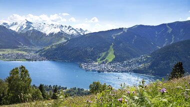 View of a shimmering lake surrounded by green hills and snow-capped mountains under a clear blue sky. | © Zell am See-Kaprun Tourismus