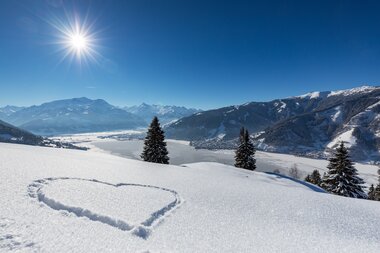 Winter landscape with snow-covered fields, trees, a heart in snow, mountains in the background, and a clear blue sky with the sun. | © Nikolaus Faistauer