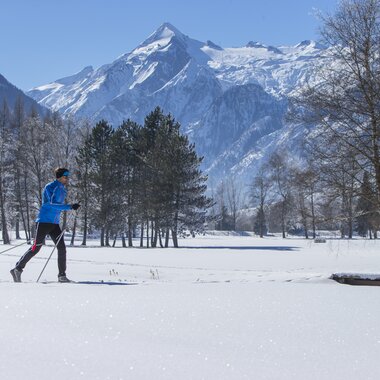 Cross-country skier in snowy landscape with mountains and trees in the background. | © Nikolaus Faistauer Photography