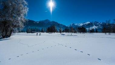 Winter landscape with snow-covered fields, trees, mountain view under clear sky, and footprints in the snow with two people in the distance. | © Zell am See-Kaprun Tourismus