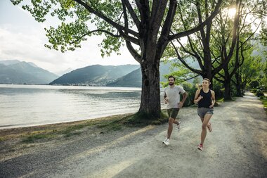 Two people running along a lake with mountains in the background, under large trees on a trail. | © Zell am See-Kaprun Tourismus