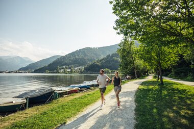 Runners jogging along a lake on a sunny day, surrounded by green trees and mountains in the background. | © Zell am See-Kaprun Tourismus