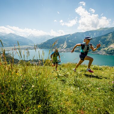 Two people trail running across a grassy meadow with flowers, mountains, a lake, and a blue sky in the background. | © Jakob Edholm