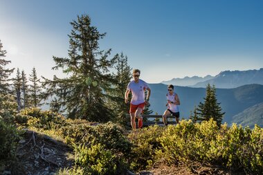 Two people trail running in the mountains with a view of the Alps in the background. | © Zell am See-Kaprun Tourismus