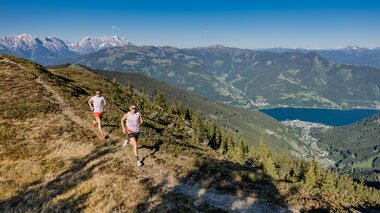 Two people trail running in the mountains with a lake and snow-capped peaks in the background. | © Zell am See-Kaprun Tourismus