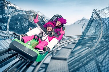 Two children are joyfully riding a toboggan on a snowy mountain slide. | © Kitzsteinhorn