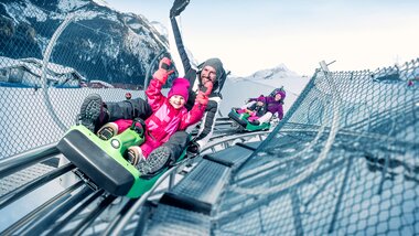 People enjoying a winter sled ride on a track with snow-covered mountains in the background. | © Kitzsteinhorn 