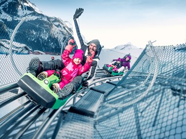 People enjoying a winter sled ride on a track with snow-covered mountains in the background. | © Kitzsteinhorn 