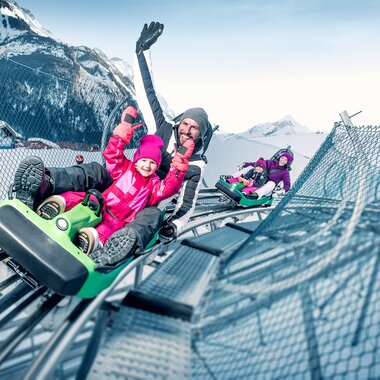 People enjoying a winter sled ride on a track with snow-covered mountains in the background. | © Kitzsteinhorn 