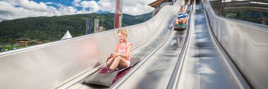 Visitors sliding down an large, silver slide at a fun park, surrounded by mountains and blue sky. | © Kitzsteinhorn