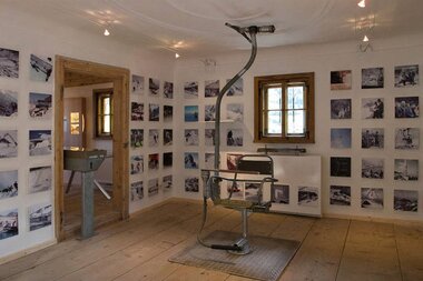 Museum exhibition room with a wall of photographs, a chair, and a window in a wooden frame. | © Kaprun Museum