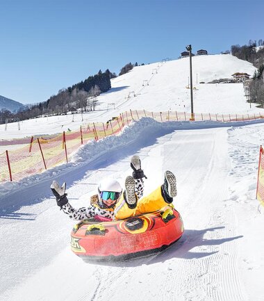 A person is snowtubing on a snowy slope with mountains and clear blue sky in the background. | © Harry Liebmann