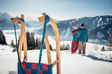 Two people with a child stand on a snowy slope enjoying the mountain view, with a sled in the foreground. | © Zell am See-Kaprun Tourismus