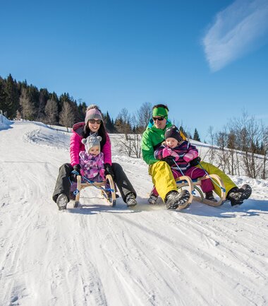 Family sledding on a snow-covered track in winter, with trees and a blue sky in the background. | © Matthias Kendler