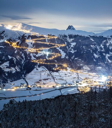 Night view of a snow-covered valley with lit sledging tracks on the mountains in the background. | © Lukas Budimaier