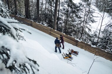 Two people sledding in a snowy forested area with sleds on the ground. | © Zell am See-Kaprun Tourismus