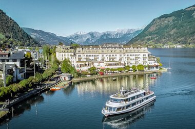 View of a hotel by the lakeside, surrounded by mountains with a yacht passing by. | © David Wöckinger