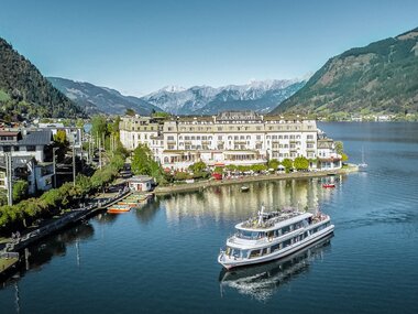 View of a hotel by the lakeside, surrounded by mountains with a yacht passing by. | © David Wöckinger