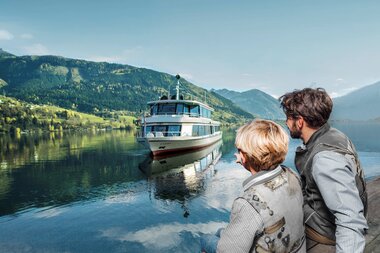 Two people watch a boat on a calm, reflective lake with mountains in the background. | © Schmittenhöhe