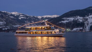 Boat decorated with festive lights on the lake with snow-covered mountains at sunset. | © Schmittenhöhebahn AG