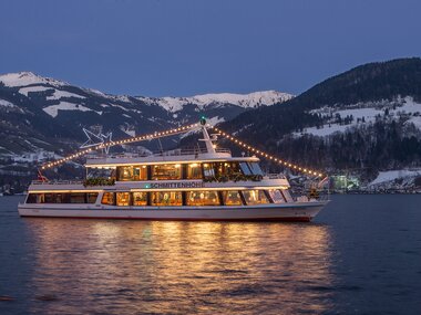 Boat decorated with festive lights on the lake with snow-covered mountains at sunset. | © Schmittenhöhebahn AG