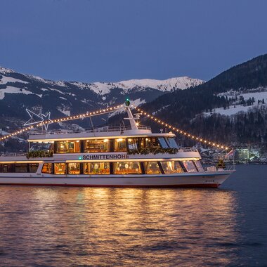 Boat decorated with festive lights on the lake with snow-covered mountains at sunset. | © Schmittenhöhebahn AG