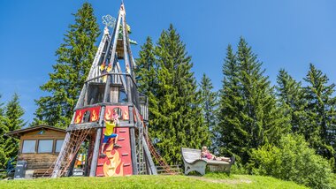 Colorful leisure facility with a large, artistically designed slide surrounded by green trees and a small wooden cabin in the background. | © Max Steinbauer