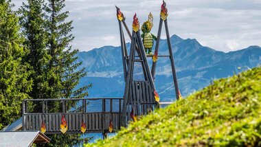 Mountain landscape with a large wooden figure on fire on a platform, surrounded by trees and mountains in the background. | © Schmittenhöhe, Max Steinbauer