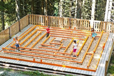 Four children are playing in a wooden maze in the forest, surrounded by trees. | © Schmittenhöhe