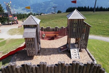 Playground with two towers, slide, and climbing wall on a field with mountain view. | © Schmittenhöhe