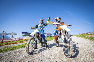 Two motocross riders in helmets, standing on their bikes, in hilly terrain on a sunny day. | © Schmittenhöhe
