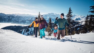 Group snowshoeing in a winter mountain landscape with snow, mountains, and pine trees in the background. | © Zell am See-Kaprun Tourismus