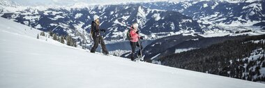 Two people snowshoeing on a snowy mountain slope with a view of mountains and a clear blue sky, heading through the winter landscape. | © Zell am See-Kaprun Tourismus