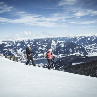 Two people snowshoeing on a snowy mountain slope with a view of mountains and a clear blue sky, heading through the winter landscape. | © Zell am See-Kaprun Tourismus